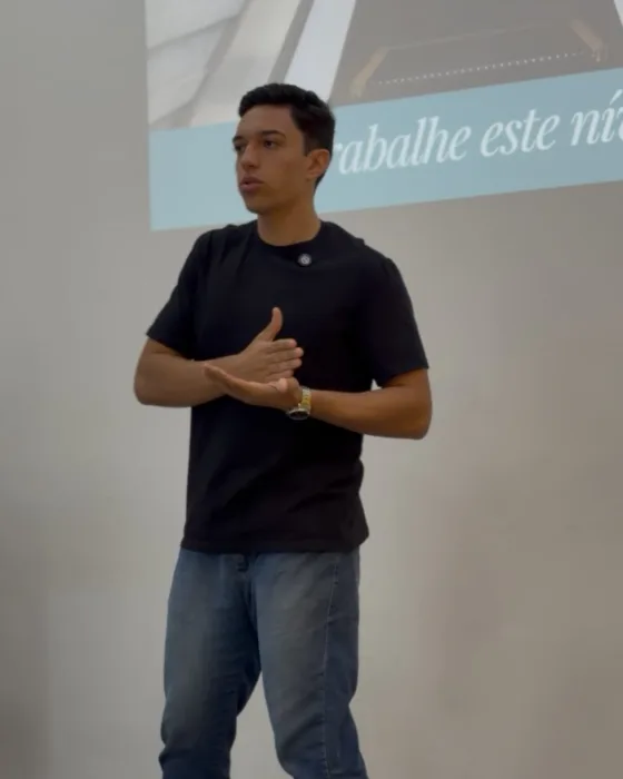Vinícius Sebriano falando em um evento, em frente a um telão